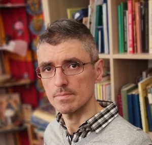 Könyvespolc előtt ülő, szemüveges férfi portréja, enyhén oldalra néz.